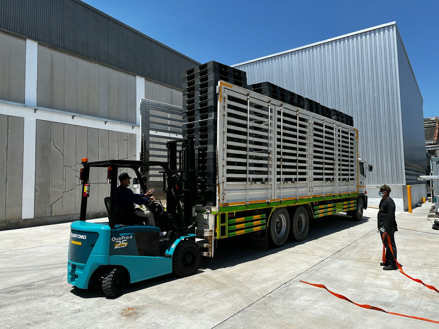 İşçi, paletleri bir kamyona yükleyen turkuaz forklifti sürüyor.